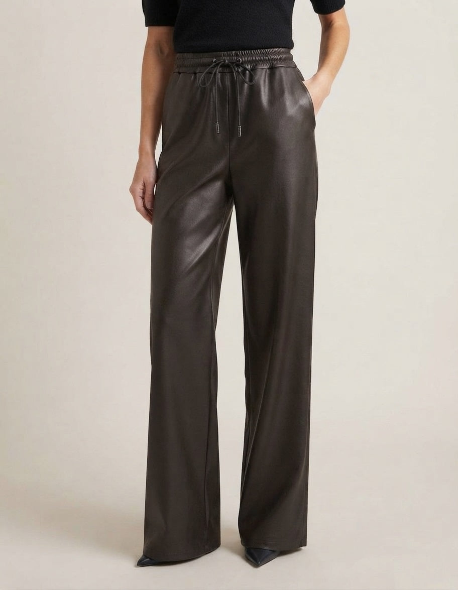 Model draagt zwarte PU‑broek met elastische tailleband en drawstring, gefotografeerd in studio tegen off‑white achtergrond. De broek valt soepel en heeft een elegante glans.