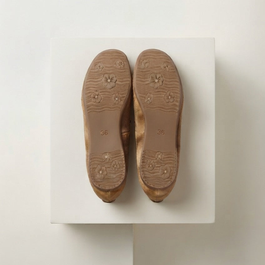 Onderkant van beige suède flats op een off‑white studio‑blok met zachte premium schaduw.