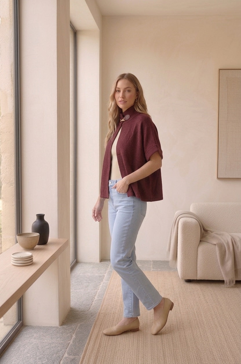 Model draagt bordeaux cape met leren gesp en beige top in een binnensetting.