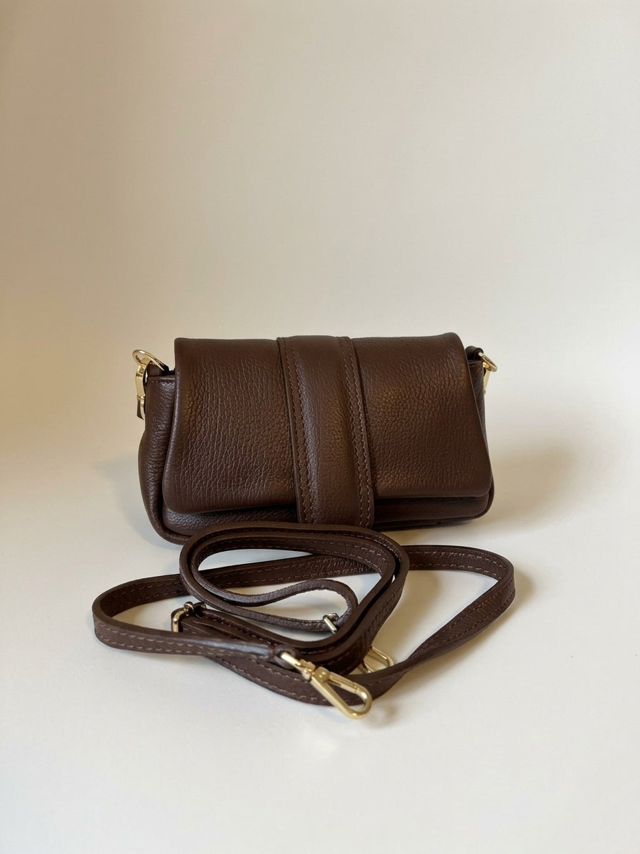 Bruine leren handtas met gold hardware en losse schouderband ervoor, high‑end neutral studio setting.
