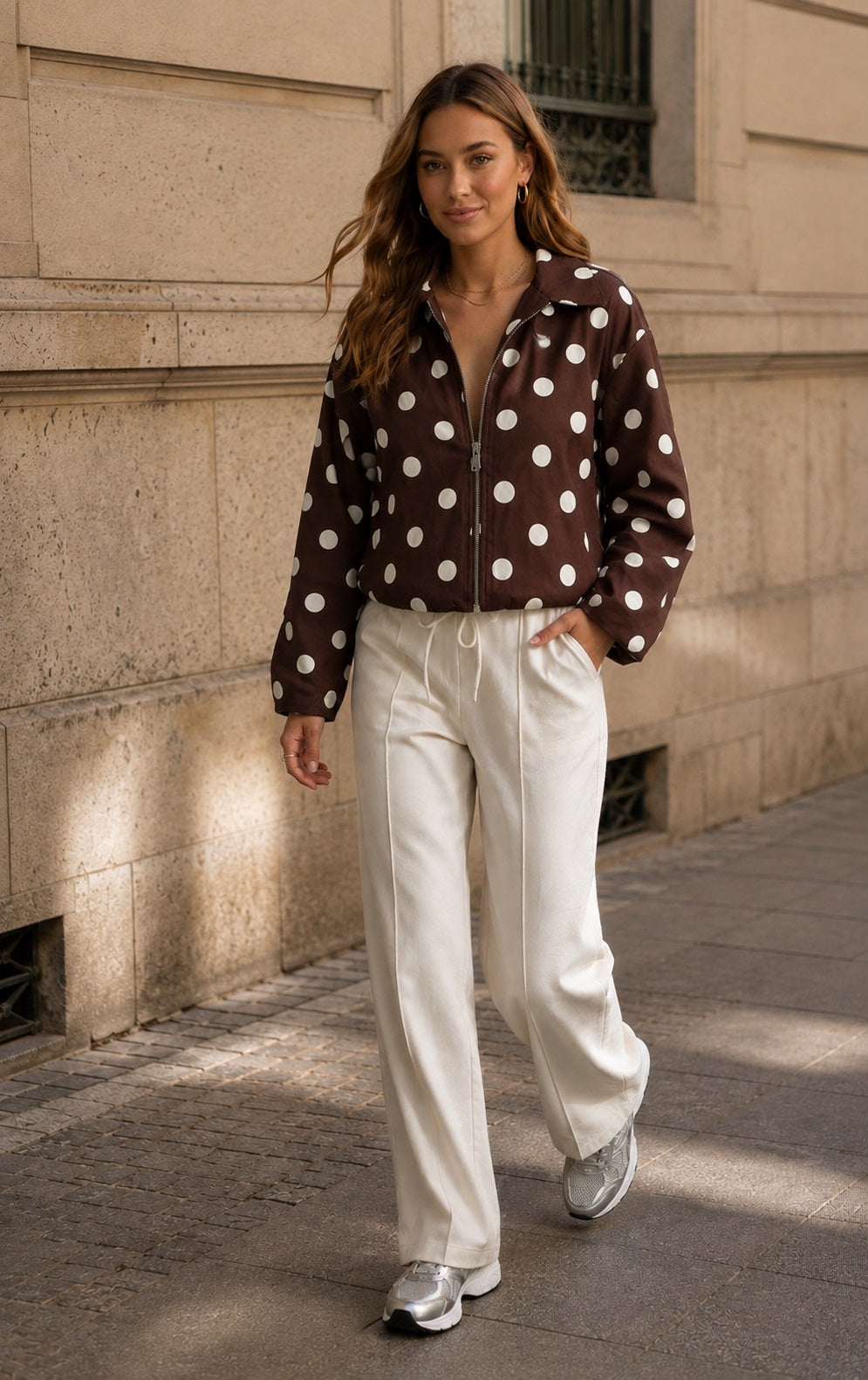 Model draagt bruine polka dot jas in streetstyle look met beige broek en sneakers, gefotografeerd in een beige urban setting.