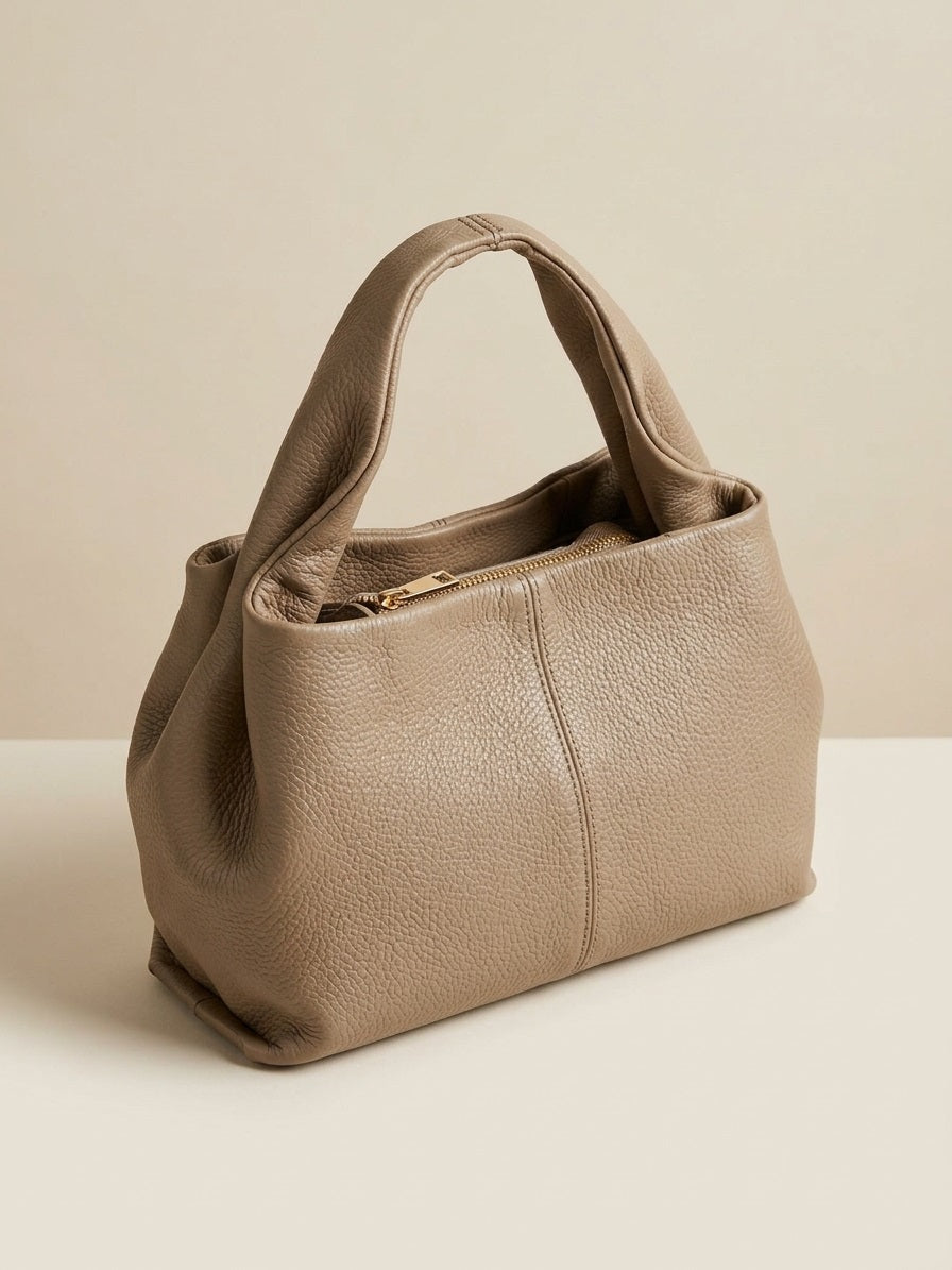 Zijaanzicht van taupe leren handtas met zichtbare naden en slouchy structuur, minimalistische studio achtergrond.