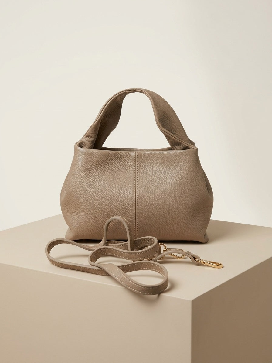 Leren schoudertas Valentina met schouderband en gold hardware naast taupe leren handtas, clean off‑white studio setting.