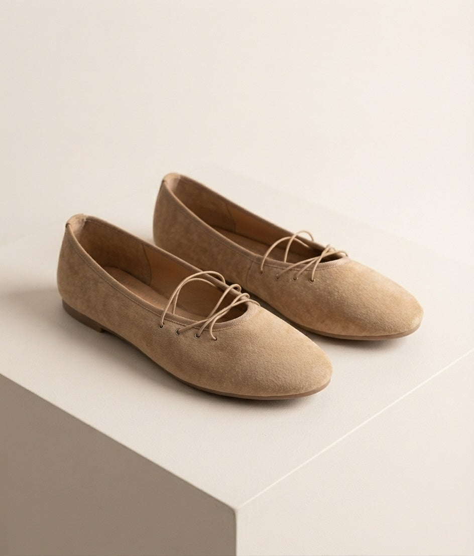 Zijaanzicht van beige suède flats met precies vier vetergaatjes en rechte veters zonder strik, geplaatst op een off‑white studio‑blok met zachte premium schaduw.