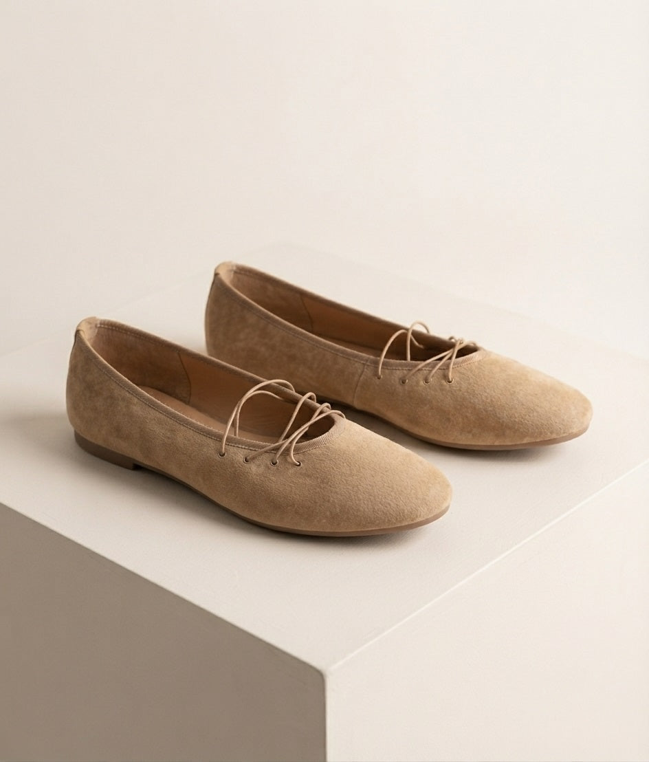 Schuin zijaanzicht van beige suède flats met precies vier vetergaatjes en rechte veters zonder strik, geplaatst op een off‑white studio‑blok met zachte premium schaduw.