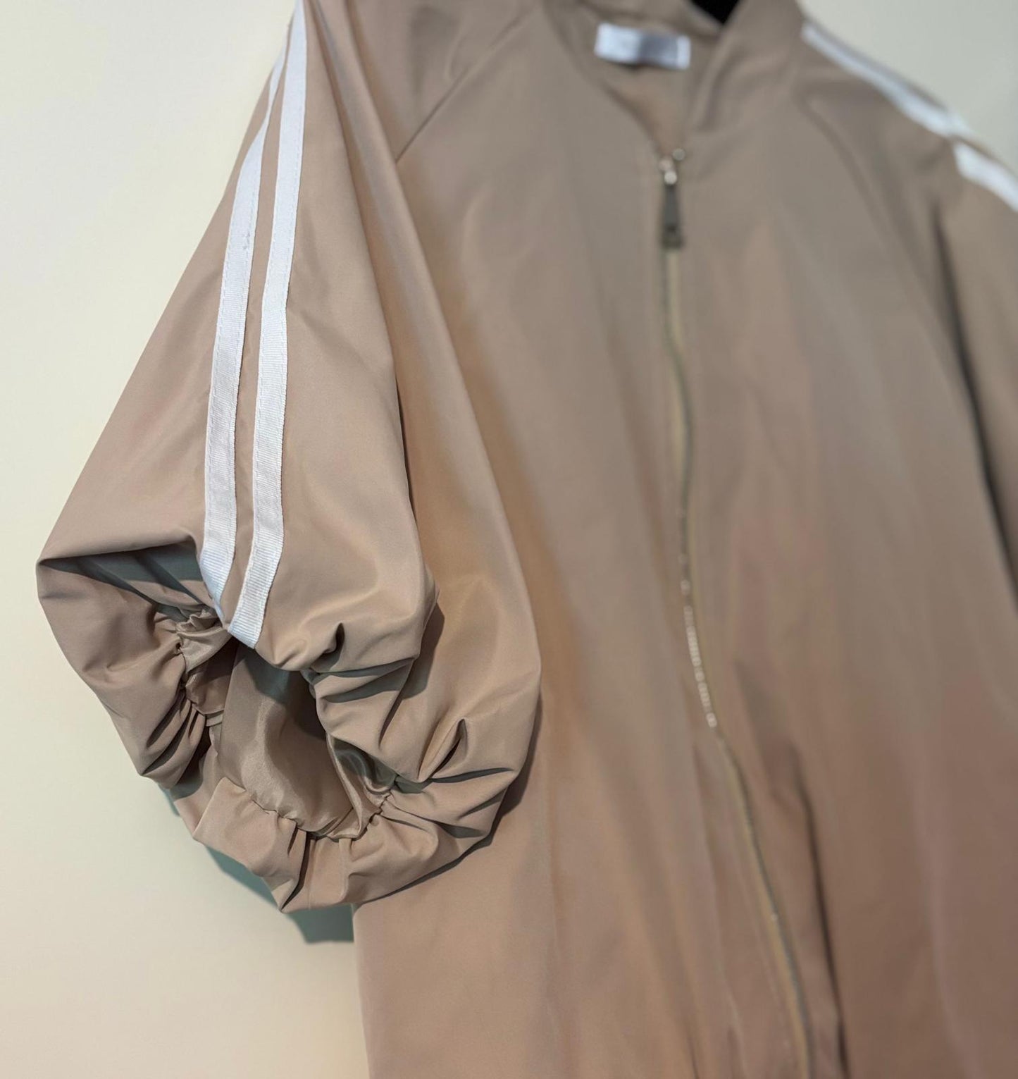 Detailfoto van de beige zip‑up track jacket met witte strepen op de mouw, hangend aan een hanger in een neutrale setting.