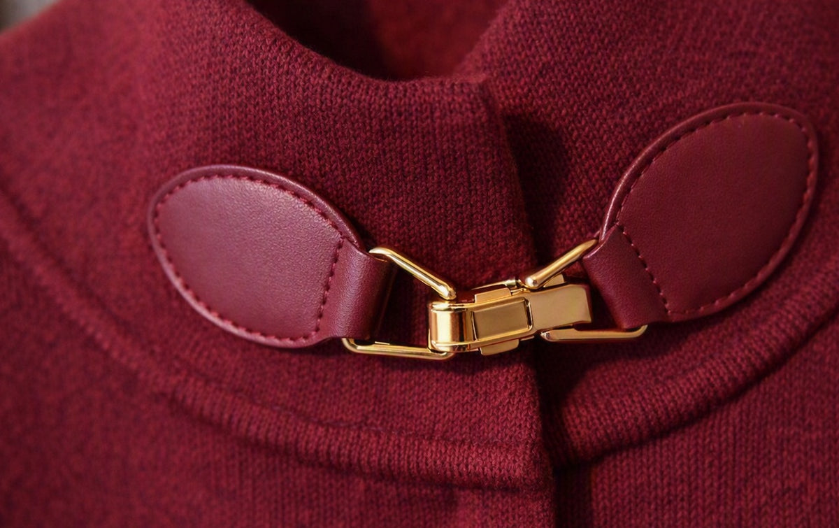 Detail van bordeaux cape met leren gesp en goudkleurige sluiting, close‑up van materiaal en afwerking.