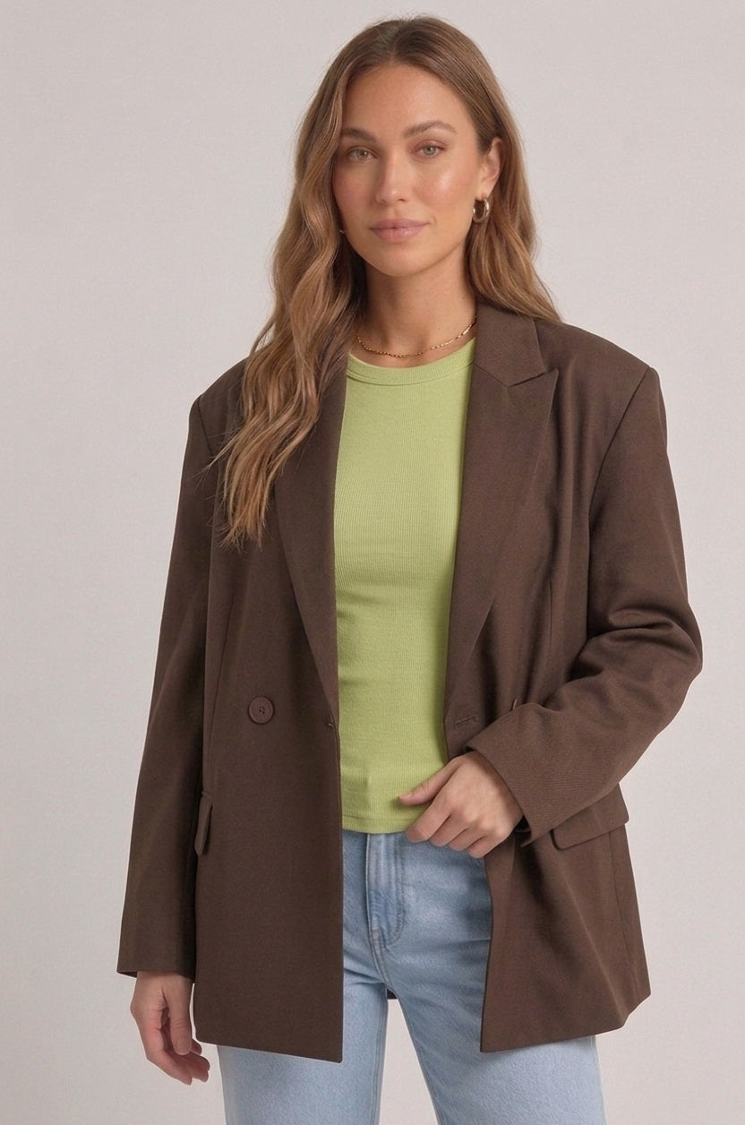 Model draagt brown oversized blazer gecombineerd met groene ribbed longsleeve in een indoor neutral setting.