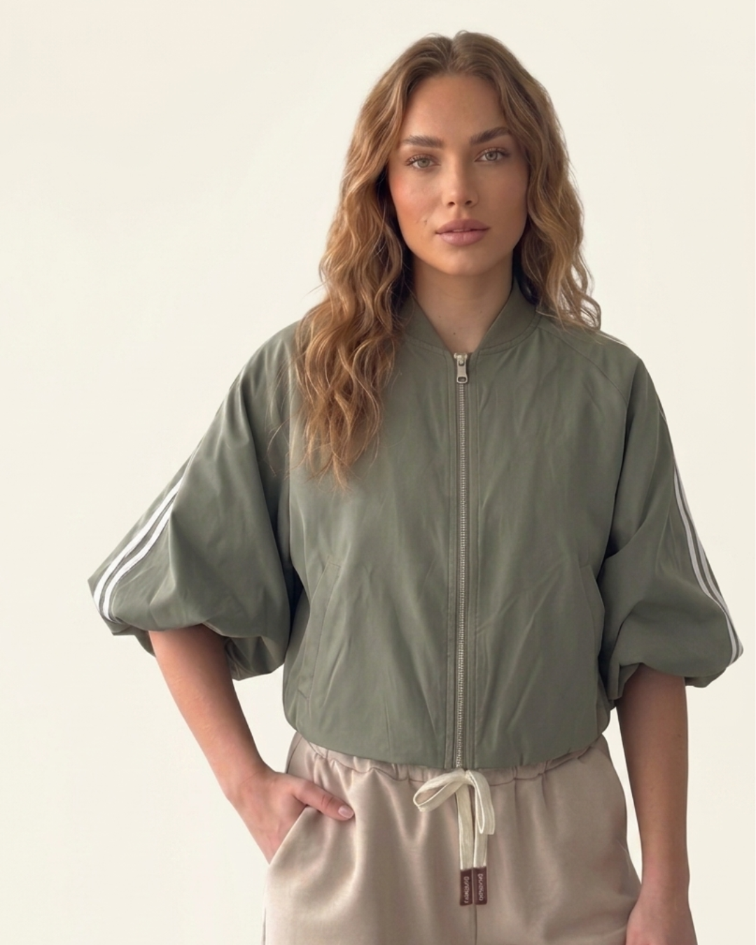 Model draagt een sage green  zip‑up track jacket met witte strepen en beige broek 
