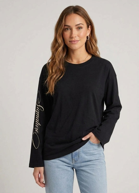 Model draagt een zwarte katoenen dames longsleeve met een ronde hals, gecombineerd met een lichtblauwe jeans. Op de rechterarm is een subtiele, sierlijke witte tekstprint zichtbaar